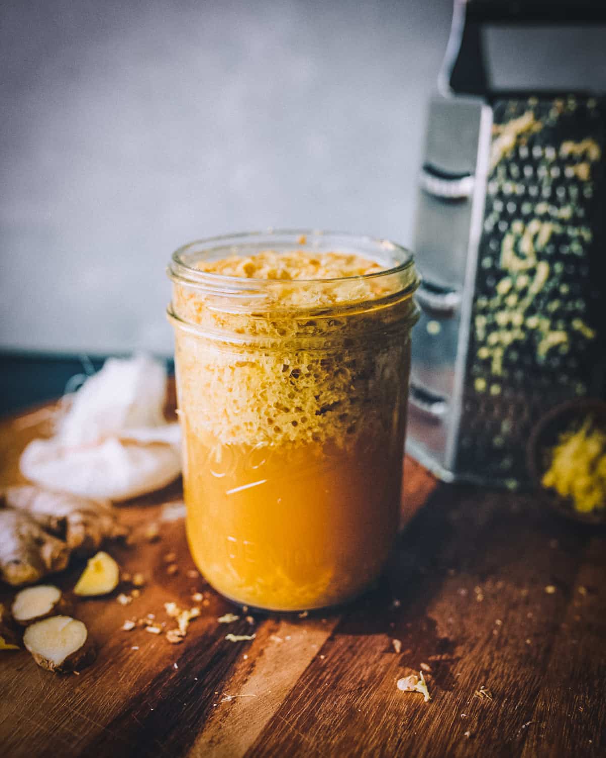A ginger bug fermenting in a jar on a dark wood surface, surrounded by fresh ginger and a grater,
