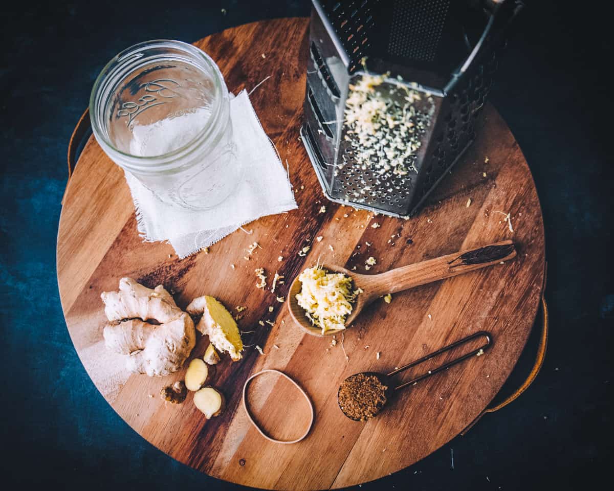 fresh ginger, a jar, a cheese grater, and spoons of ginger and sugar on a round wooden board