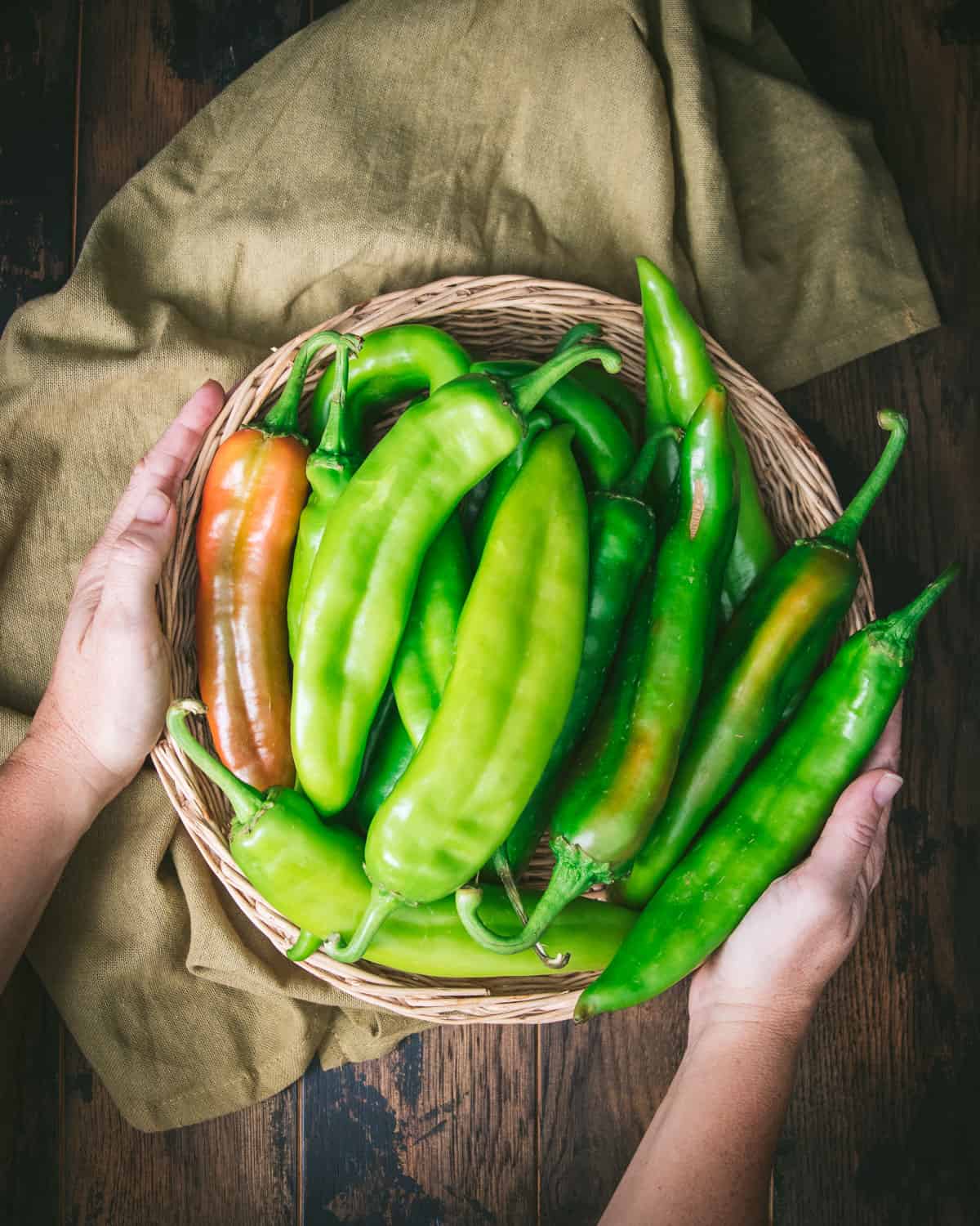 Fresh green chile peppers piled in a basket held by two hands, with an olive green cloth. 