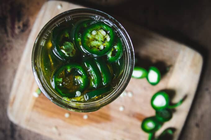 Top view of jar filled with sliced jalapeños and honey, with a wood cutting board and sliced tops of fresh jalapeños in background.