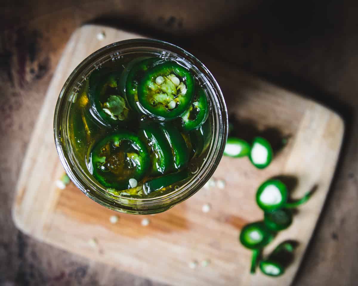 Top view of jar filled with sliced jalapeños and honey, with a wood cutting board and sliced tops of fresh jalapeños in background. 