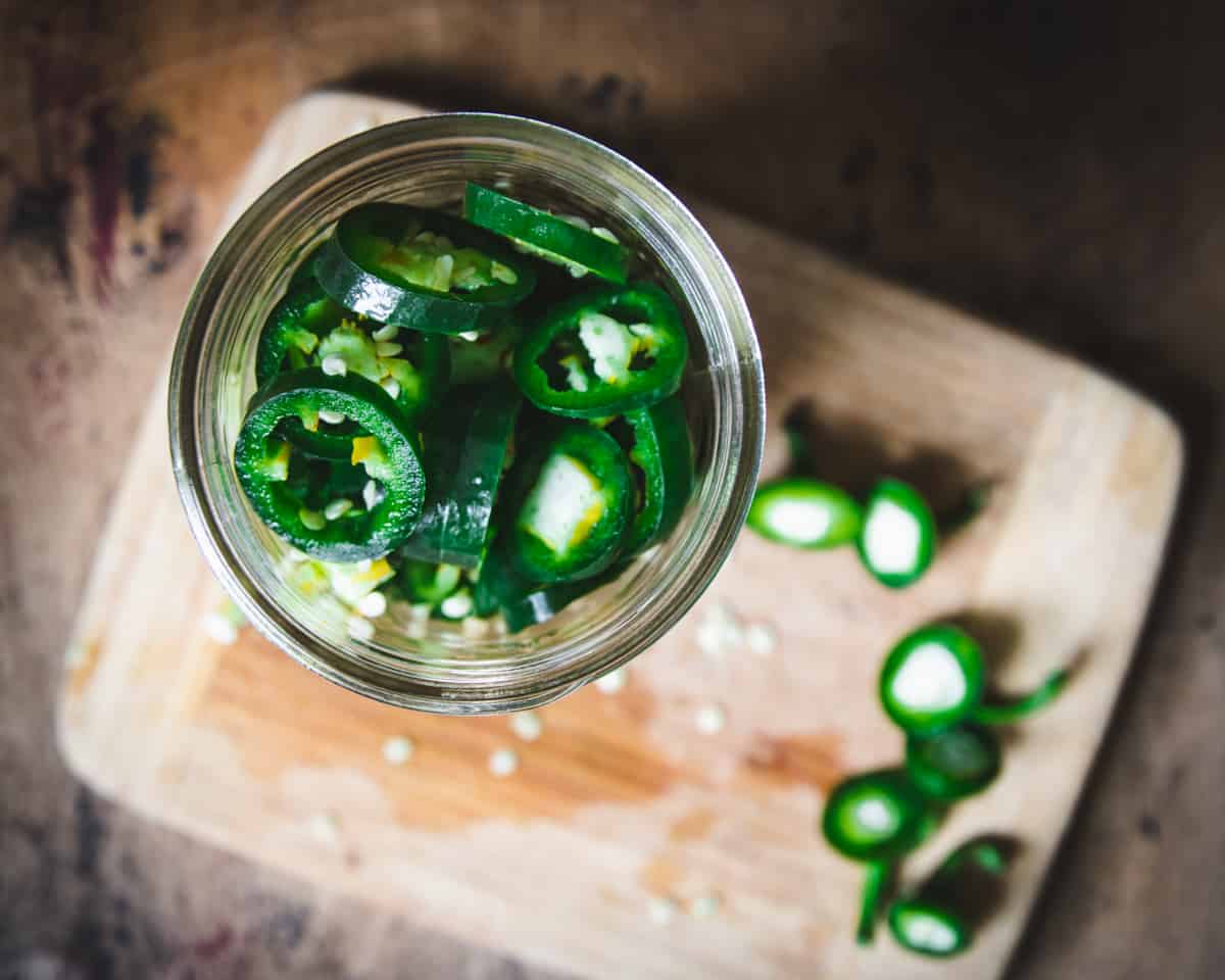 Sliced jalapeños in a jar, top view. Wooden cutting board with sliced jalapeño tops in background.