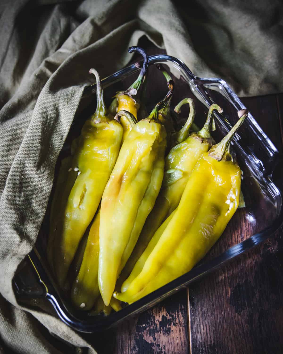 Roasted green hatch peppers piled in a square glass baking dish with an olive green fabric background.