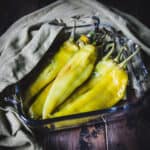 Angled side view of whole roasted green Chile peppers in a square roasting dish, with an olive green cloth in the background.