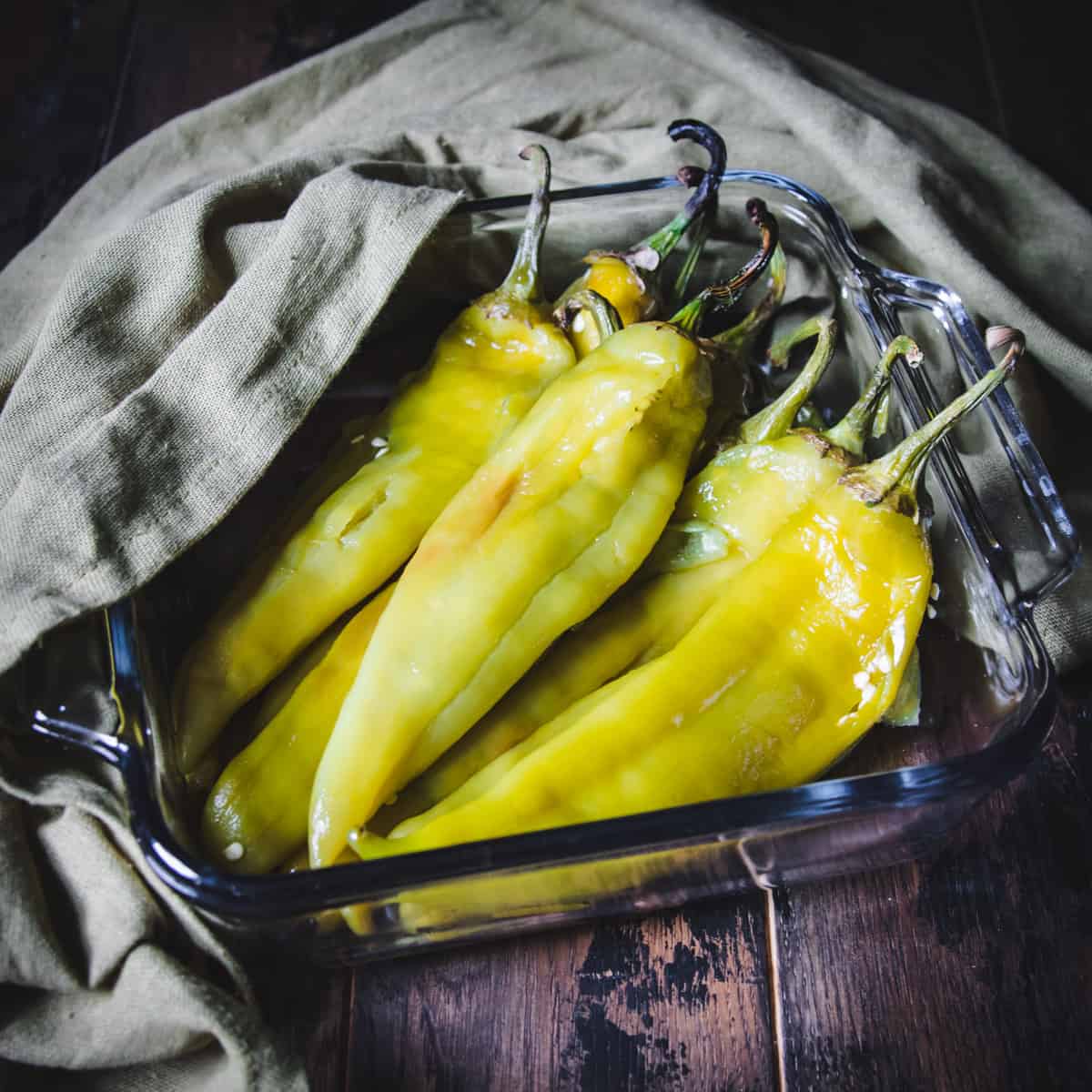 Angled side view of whole roasted green chile peppers in a square roasting dish, with an olive green cloth in the background. 