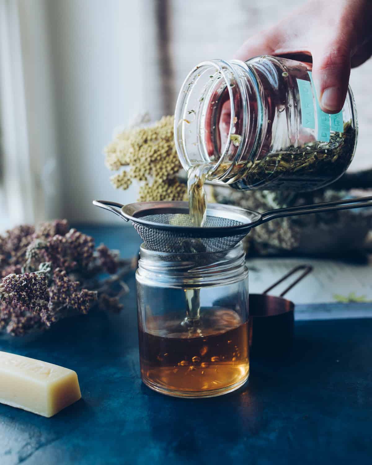 Jar of yarrow flower infused tea water, pouring into a strainer held by a clear jar. The jar is half full of strained yarrow water. On a dark blue counter top, with dried yarrow flowers and a stick of beeswax in the background. 