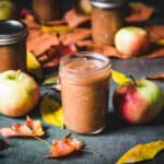 Apple butter in a jar surrounded by apples and fall leaves.