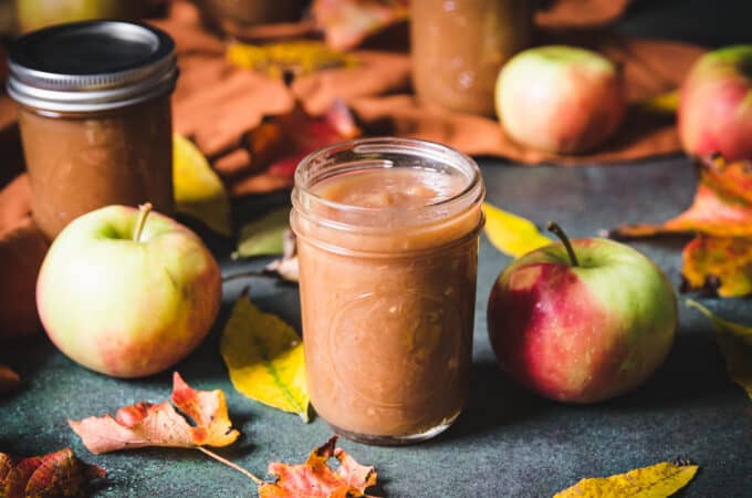 Apple butter in a jar surrounded by apples and fall leaves.