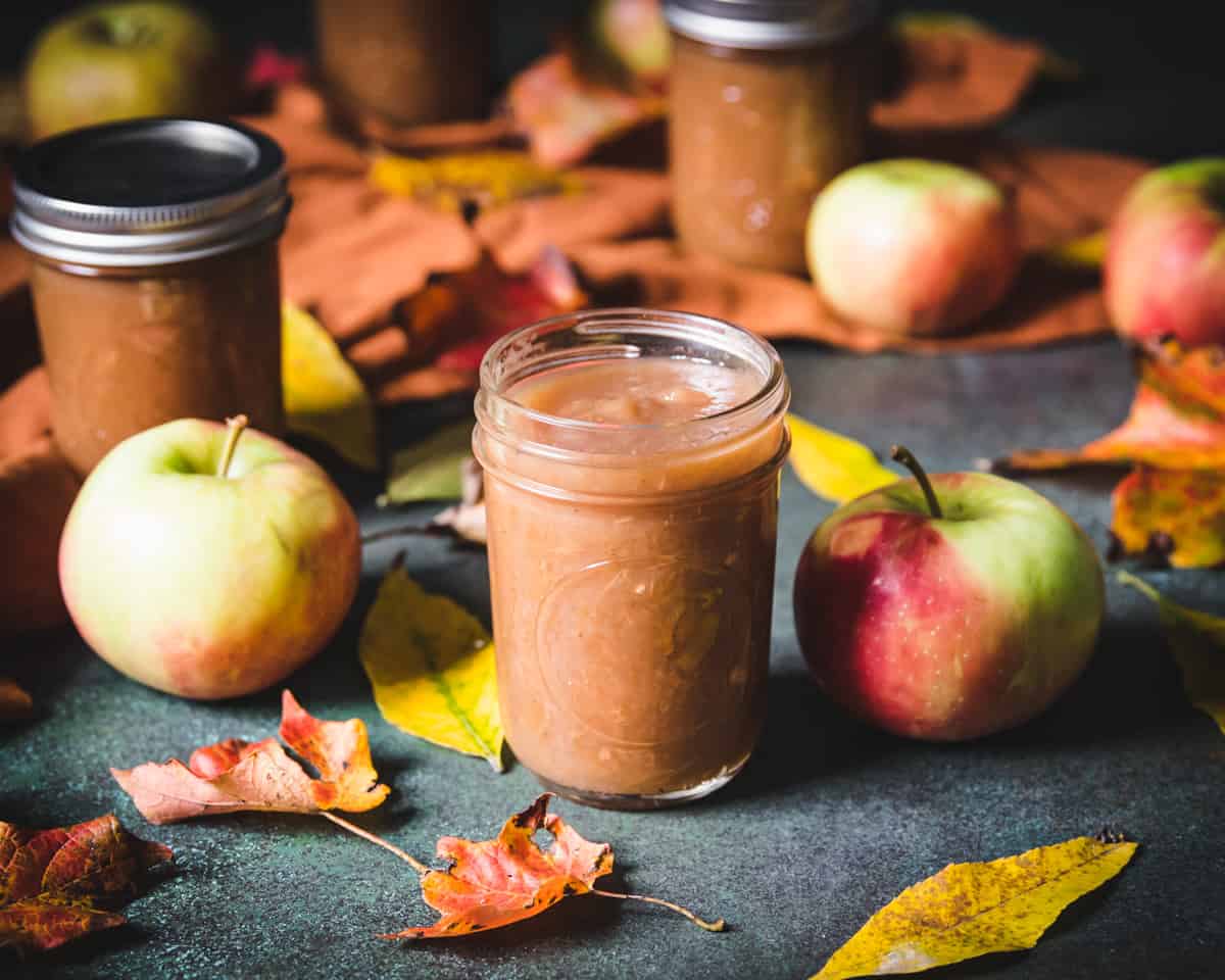 Apple butter in a jar surrounded by apples and fall leaves. 