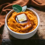 Butternut mash in a white bowl with a pat of butter and maple syrup, with fresh sage leaves. On a dark wood surface surrounded with a dark orange cloth.