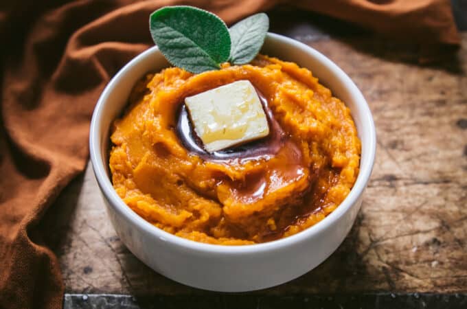 Butternut mash in a white bowl with a pat of butter and maple syrup, with fresh sage leaves. On a dark wood surface surrounded with a dark orange cloth.