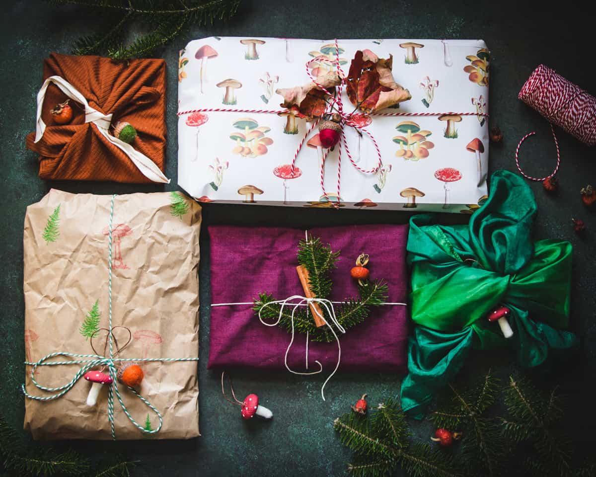 2 gifts wrapped with cloth and tied on top, 3 wrapped in paper: one that is brown and stamped with trees, one with mushroom print, one a deep magenta, all have pine sprigs, dried leaves, and felted mushrooms on top tied with natural twine. 