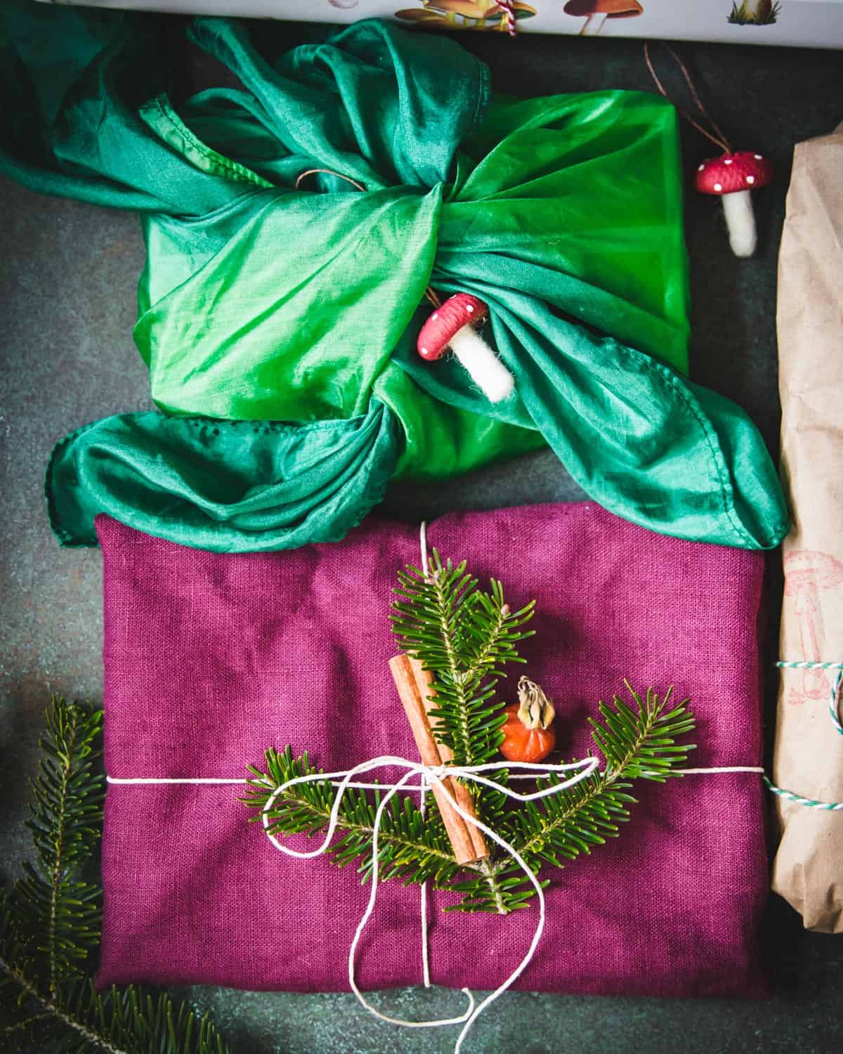 Dark grey background with 2 presents wrapped in fabric, one a deep magenta with white twine and a green pine tree twig with a cinnamon stick on top, and the other an ombre green silk fabric that is tied around the present, with felted wool mushrooms adorning. 