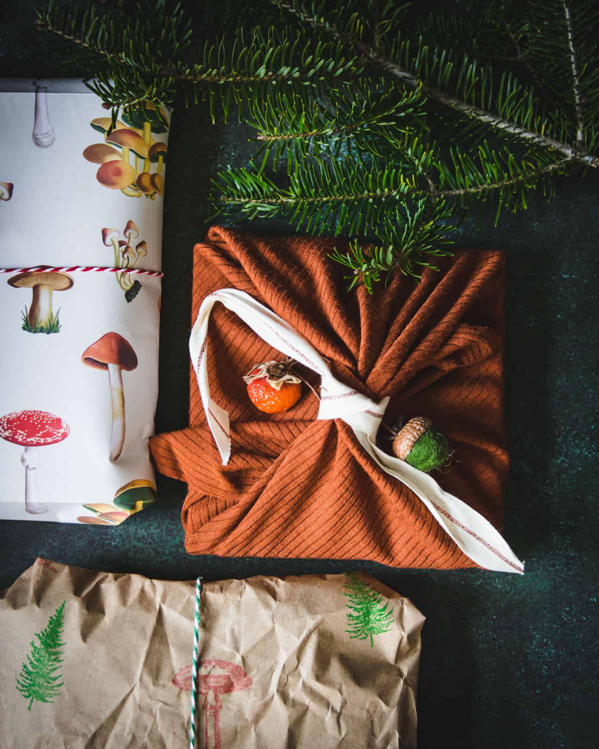 Dark background with a square present wrapped with rust colored fabric, tied with a white ribbon. Also partially shows 2 presents wrapped in paper, one with mushroom print, and one natural brown with tree and mushroom stamps on them. At top is a dark green branch of a Christmas tree. 
