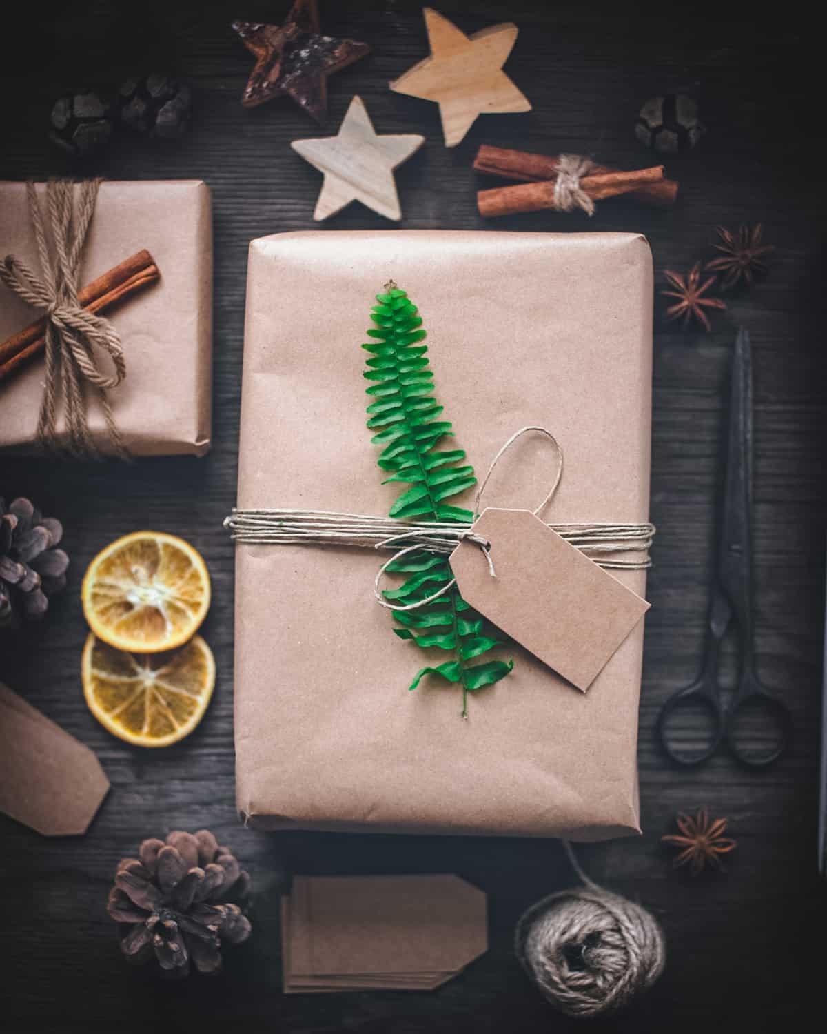 Dark wood background, with one small present wrapped in plain brown paper that is tied with natural twine and a cinnamon stick on top, and a second larger rectangle present wrapped in plain brown paper with a natural twine and a small brown tag, topped with a green fern leaf. Presents are surrounded by: dried orange slices, pine cones, black rustic scissors, a ball of twine, a star anise, brown gift tags, cinnamon sticks, and 3 wooden star cut outs. 