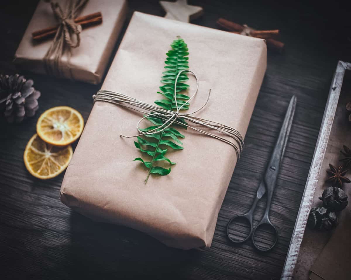 A dark wood background with 2 presents wrapped in plain brown paper, one tied with twine topped with cinnamon sticks, the bigger one in front tied with thin twine topped with a fern leaf. Surrounding the presents are: 2 dried orange slices, pine cones, black rustic scissors, and a wooden star cut out. 