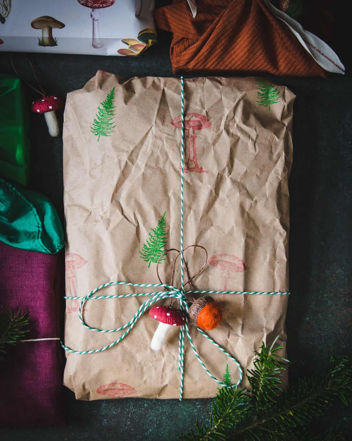 Dark background, with one present wrapped in crinkly brown paper stamped with green trees and red mushrooms, with a green and white twine bow, and a felted wool mushroom and acorn on top. 