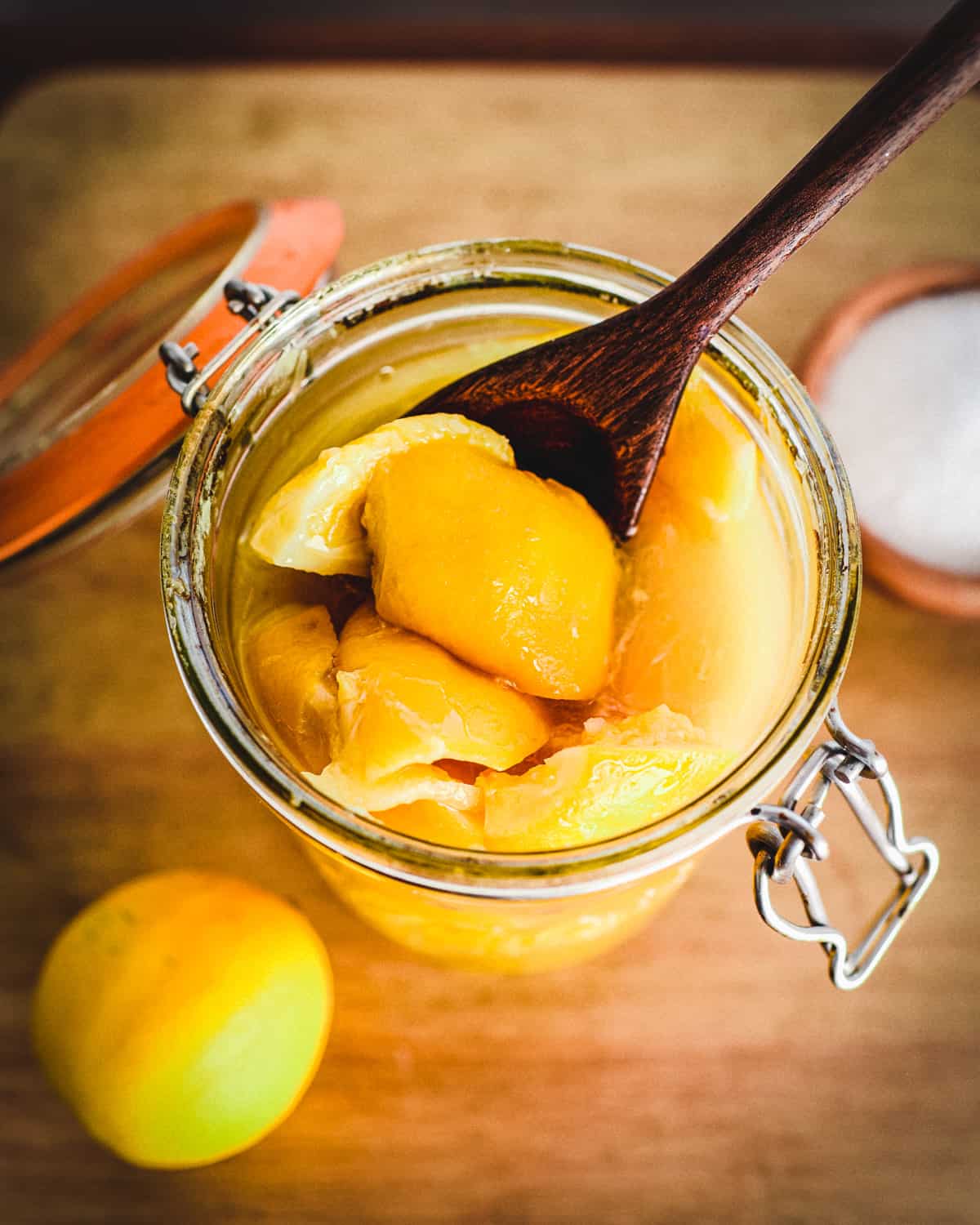 An open jar of fermented lemons with a wooden spoon holding one up into better view, on a wooden cutting board with a whole lemon on the bottom left of the background. 