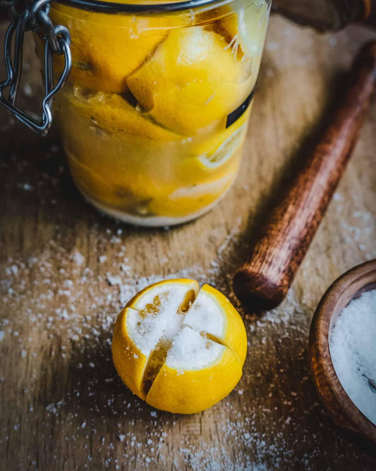 A jar filled with fermenting lemons, on a wood cutting board with a wooden muddler and a lemon outside the jar that is sliced and salted. 