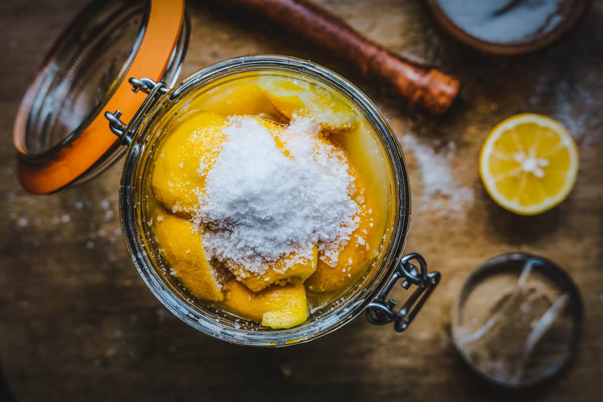 An open jar of lemons in brine with a small mountain of salt topping the lemons. In background is a wooden cutting board, a lemon sliced in half, and a wooden muddler. 