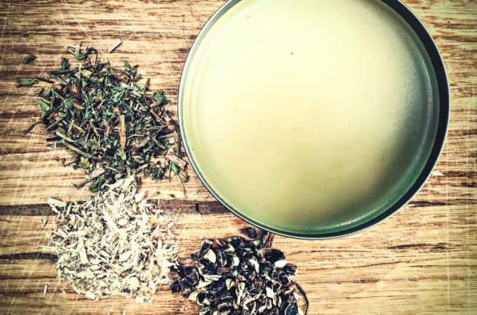 A tin of diaper rash salve on a wooden surface with piles of dried herbs surrounding.