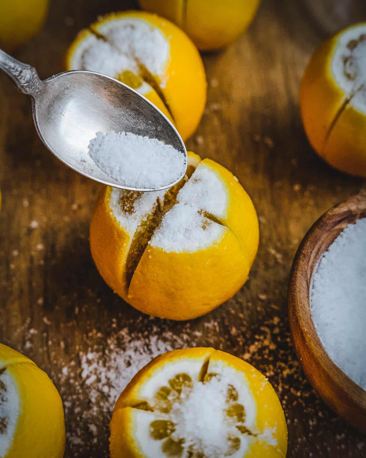 Close up of a lemon standing on end sliced in quarters with a spoon pouring salt into the opening, on a wood cutting board. 
