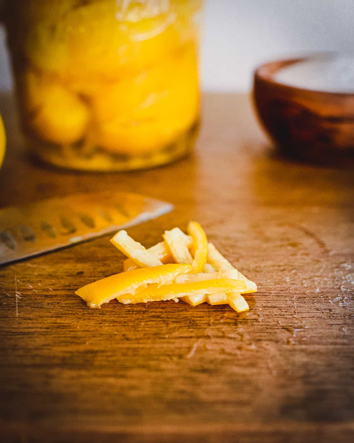 Fermented lemon peels, sliced into little strips, resting on a wood cutting board, with a sharp knife, a jar of fermented lemons, and a bowl of salt in the background. 