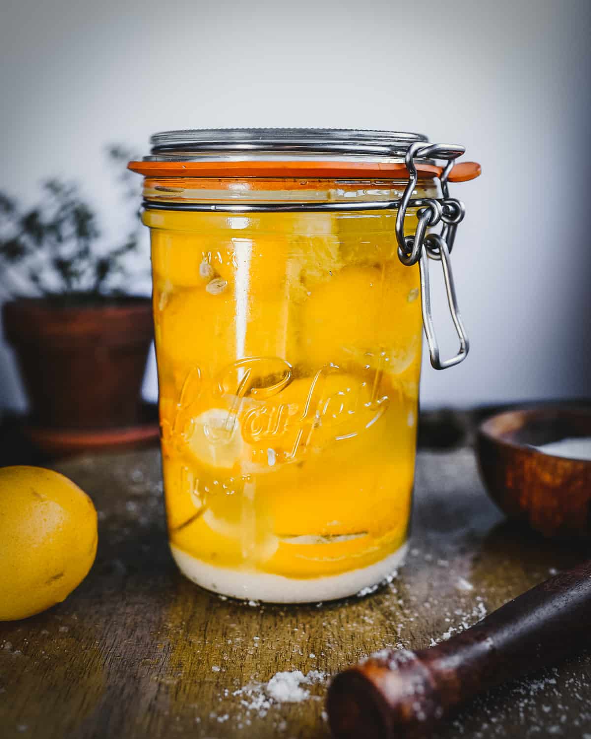 A closed jar filled with lemons submerged in salt brine, resting on a wooden cutting board, surrounded by a lemon, a wooden muddler, and a wooden bowl of salt. 