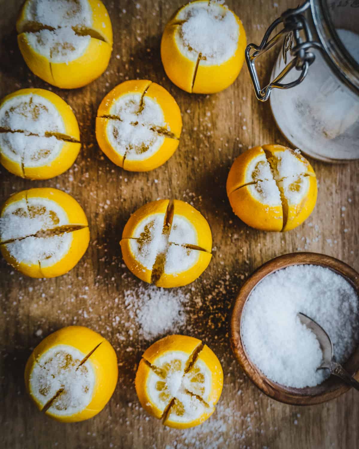 Lemons quartered, standing on connected ends and sprinkled with salt, an open jar with salt at the bottom on right, below that is a wooden bowl of salt with a spoon. All resting on a wooden cutting board. 
