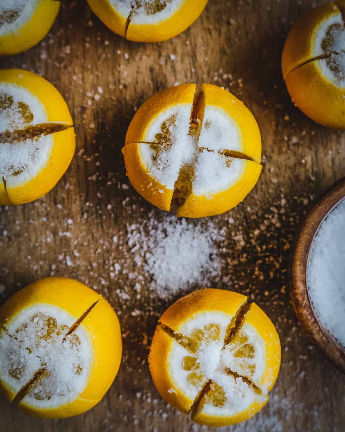 Lemons standing upright, sliced in quarters except for the bottom half inch keeping them connected at the bottoms. All are sprinkled with salt, and resting on a wooden cutting board. 