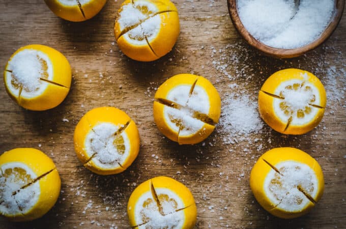 Lemons standing on end sliced in quarters but connected at bottom, all sprinkled with salt. A wooden bowl of salt with a spoon is at top right corner. All resting on a wooden cutting board.