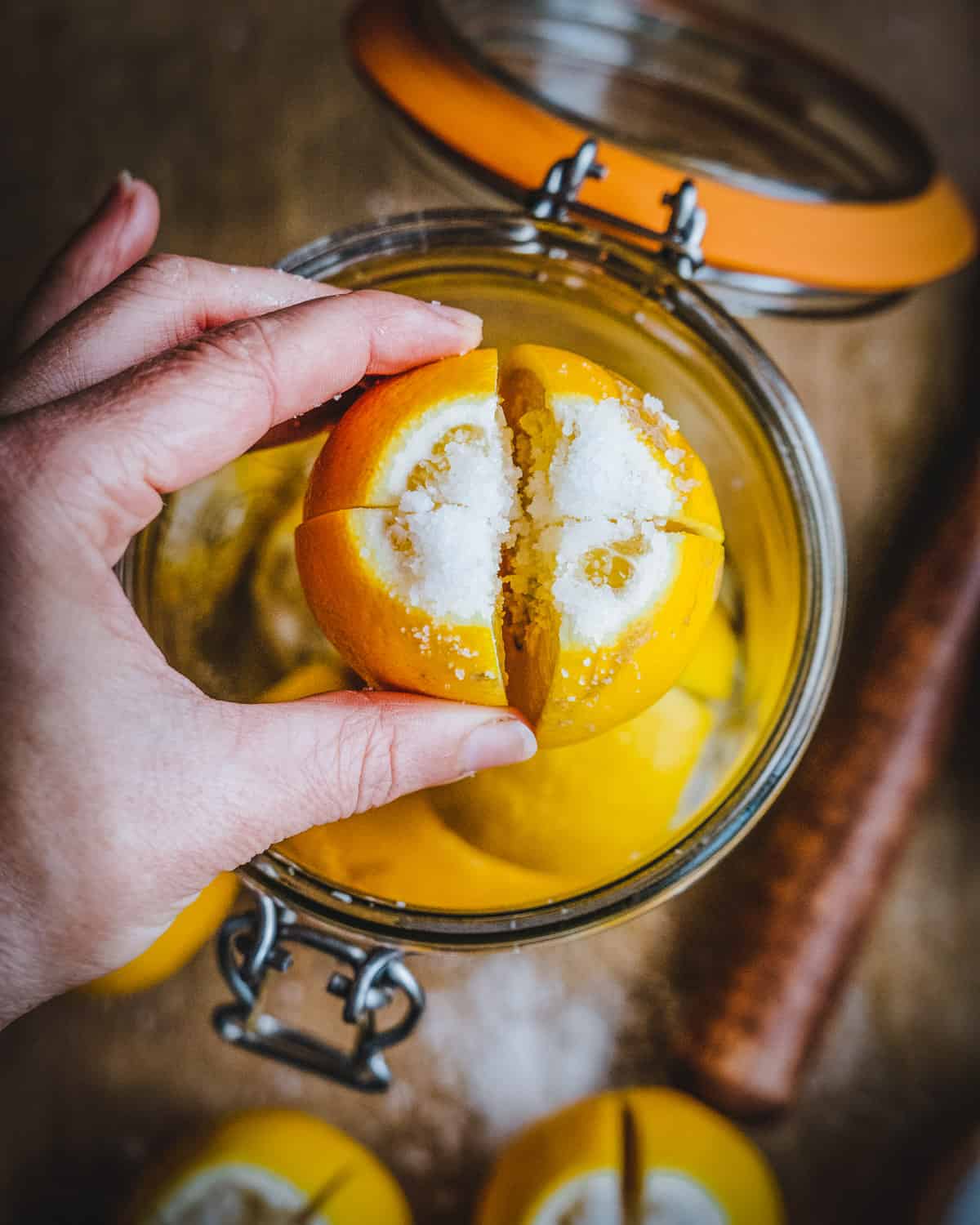 A top view of an open jar of lemons, with a hand holding a quartered lemon sprinkled with salt in the foreground. The background has a wood cutting board, several other lemons, and a wooden muddler. 