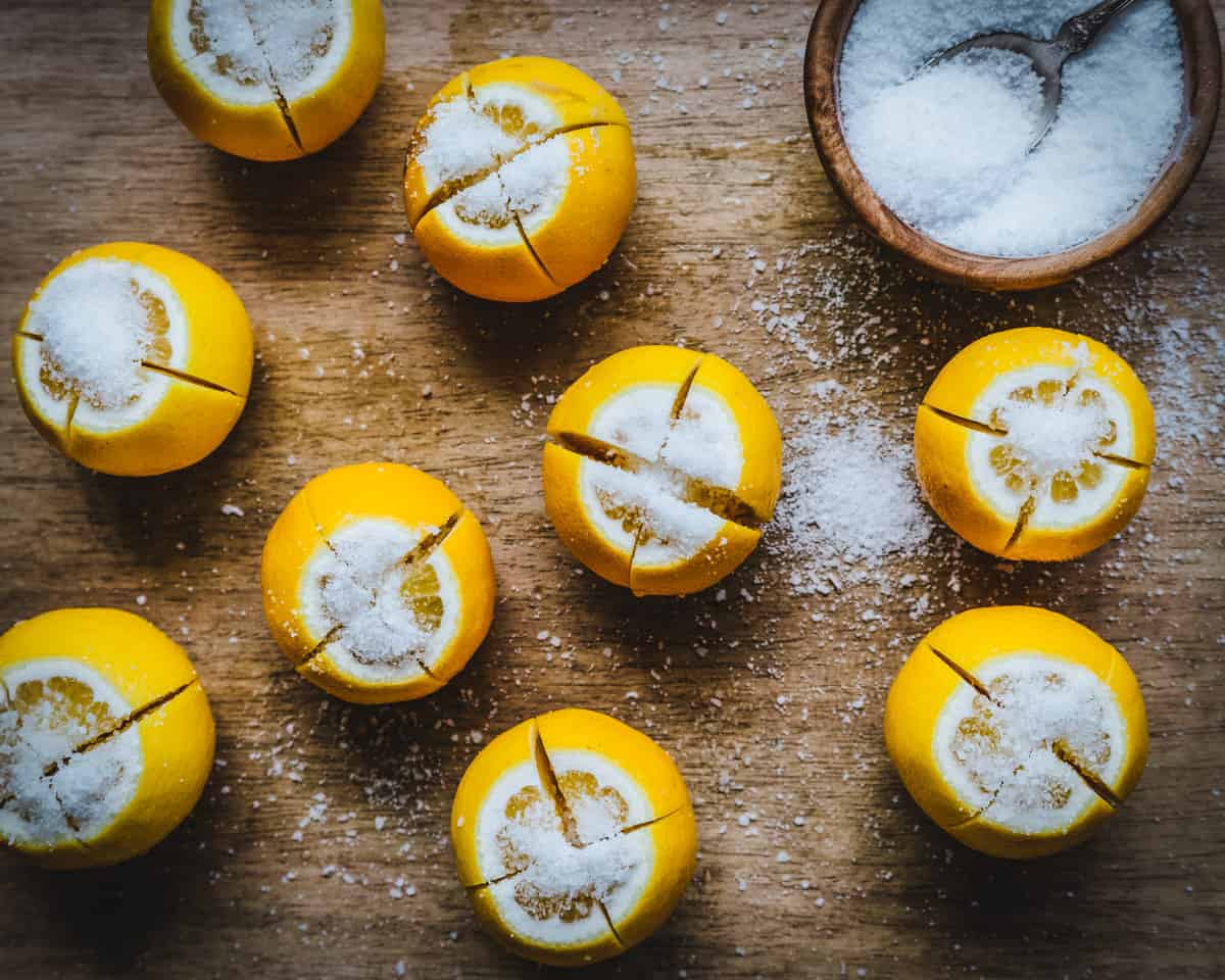 A wooden cutting board with several lemons standing on end, sliced and sprinkled with salt. A wooden bowl with a spoon and filled with salt are in the upper right corner. 