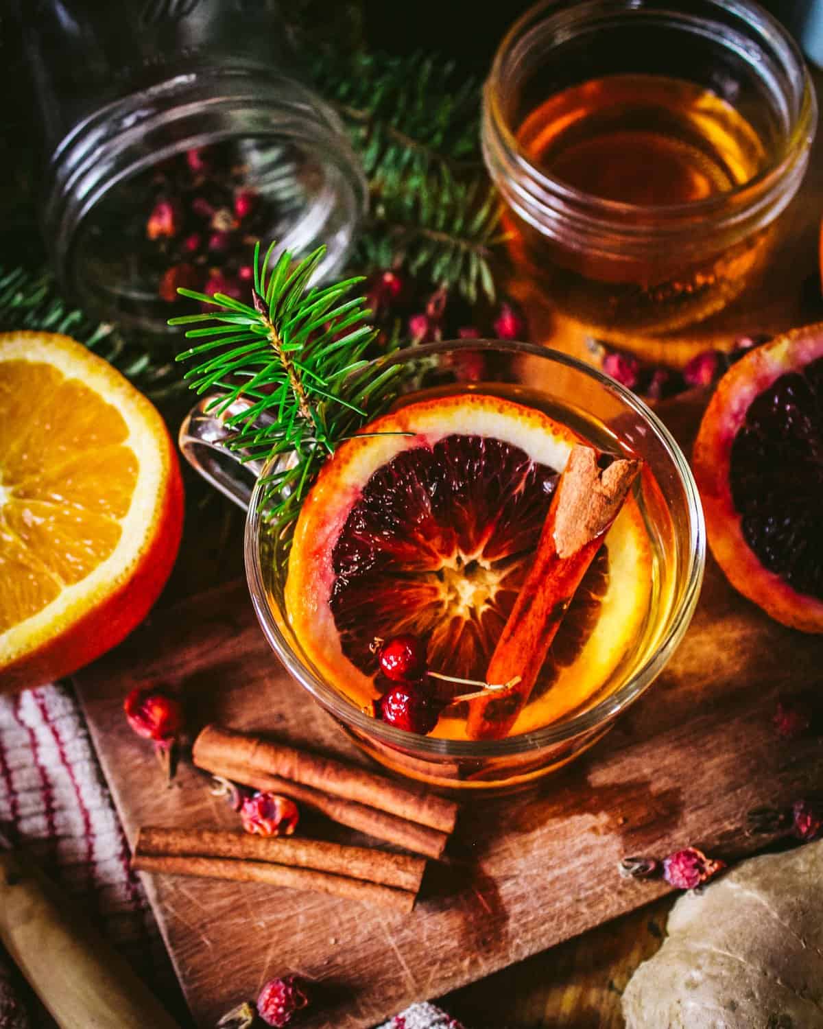 A hot toddy in a clear mug with conifer needles, cinnamon sticks, rosehips, and oranges surrounding. 