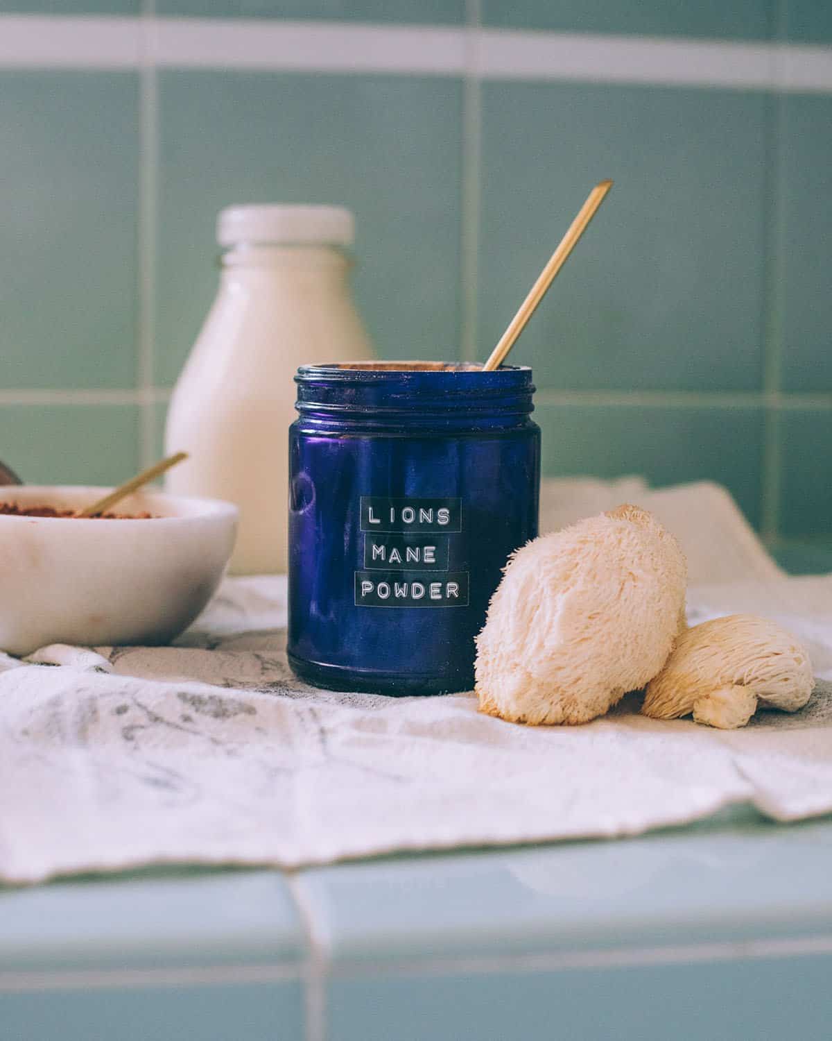 Lion's mane powder in a dark blue labeled jar with a gold spoon in it, with young lion's mane mushrooms to the side. On a white cloth sitting on a green tile counter. 