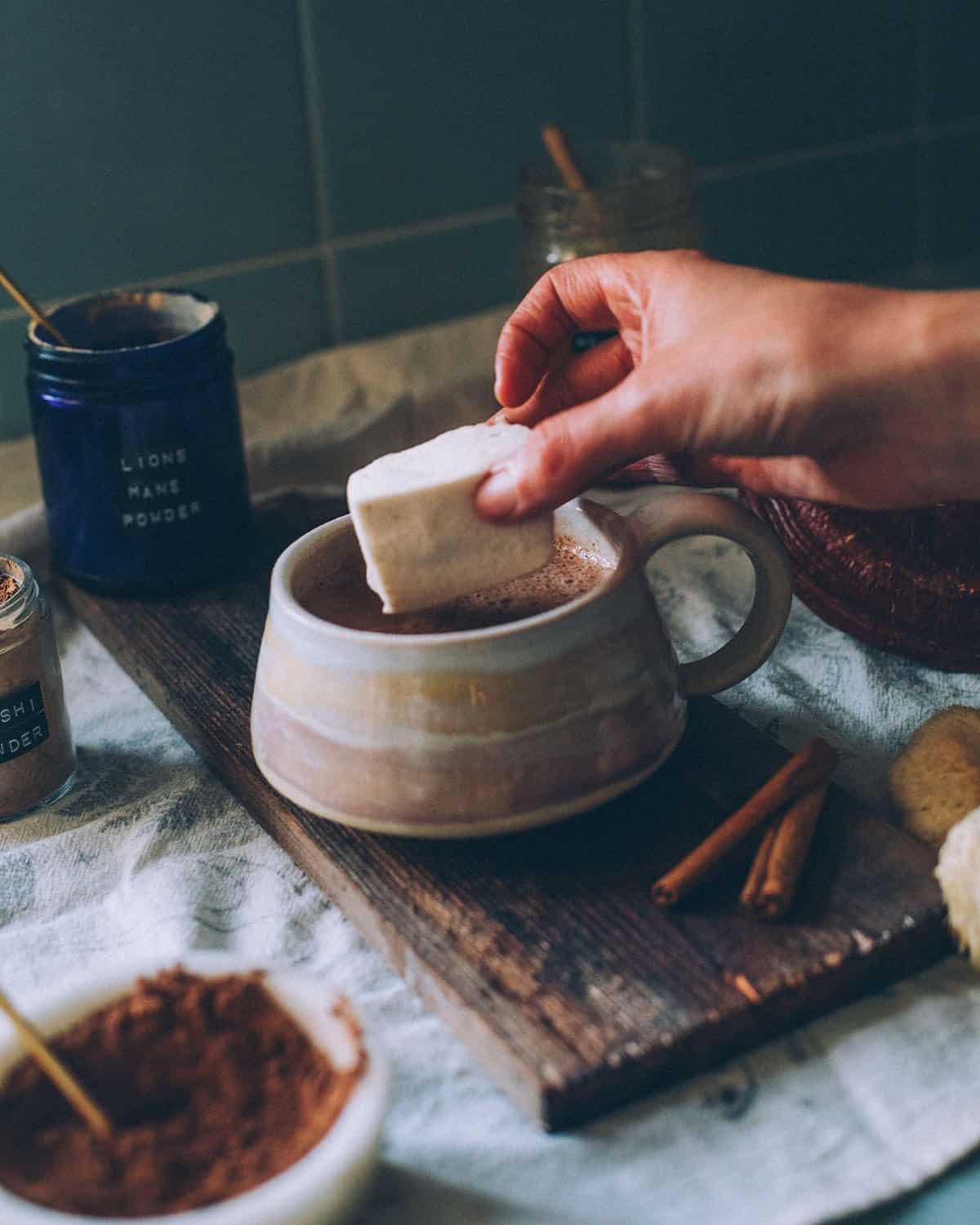 a hand putting a homemade marshmallow into a mug of mushroom hot chocolate