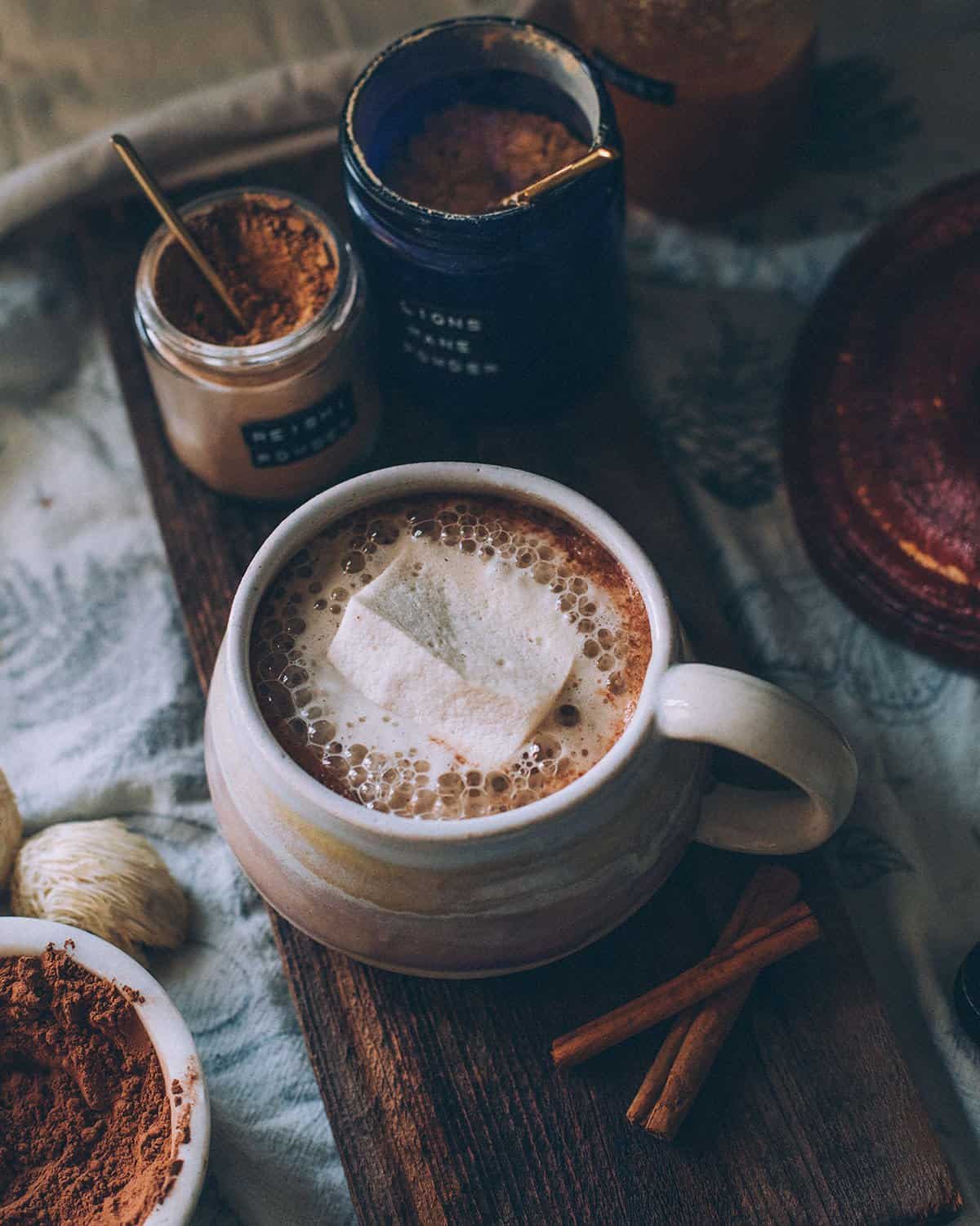 a mug of mushroom hot cocoa with a marshmallow and jars of mushroom powder