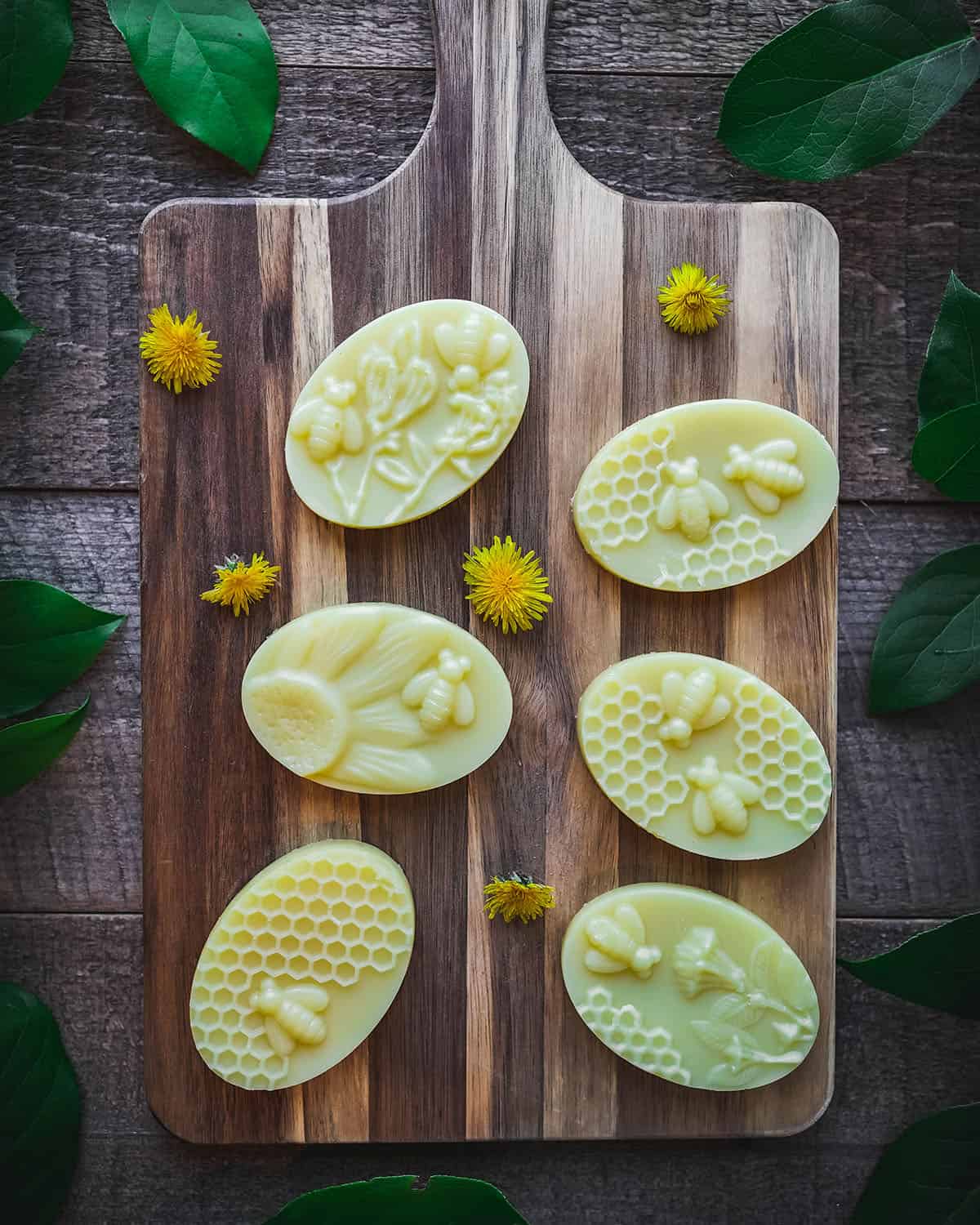 Finished dandelion lotion bars on a wood cutting board surrounded by fresh dandelions. 