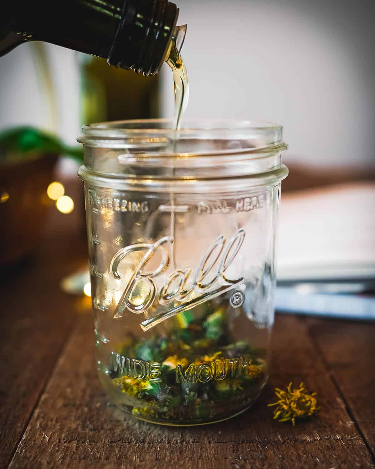 A glass jar filled partway with dried dandelions, and oil pouring into the jar. 