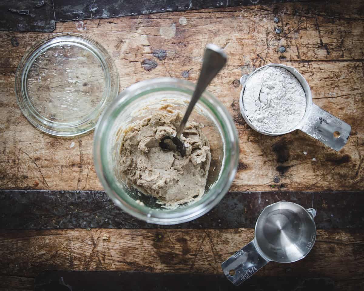 A top view of a jar with a spoon and newly mixed flour and water, to the side a measuring cup full of flour. 