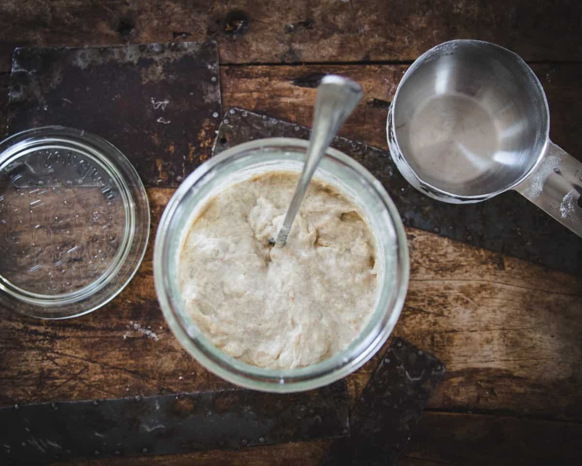 Top view of sourdough starter that is growing and stirred with a spoon. A measuring cup and glass lid sit to the side. 