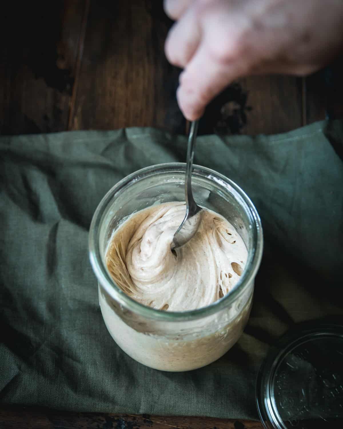 A hand stirring a sourdough starter resting on a forest green natural fabric.