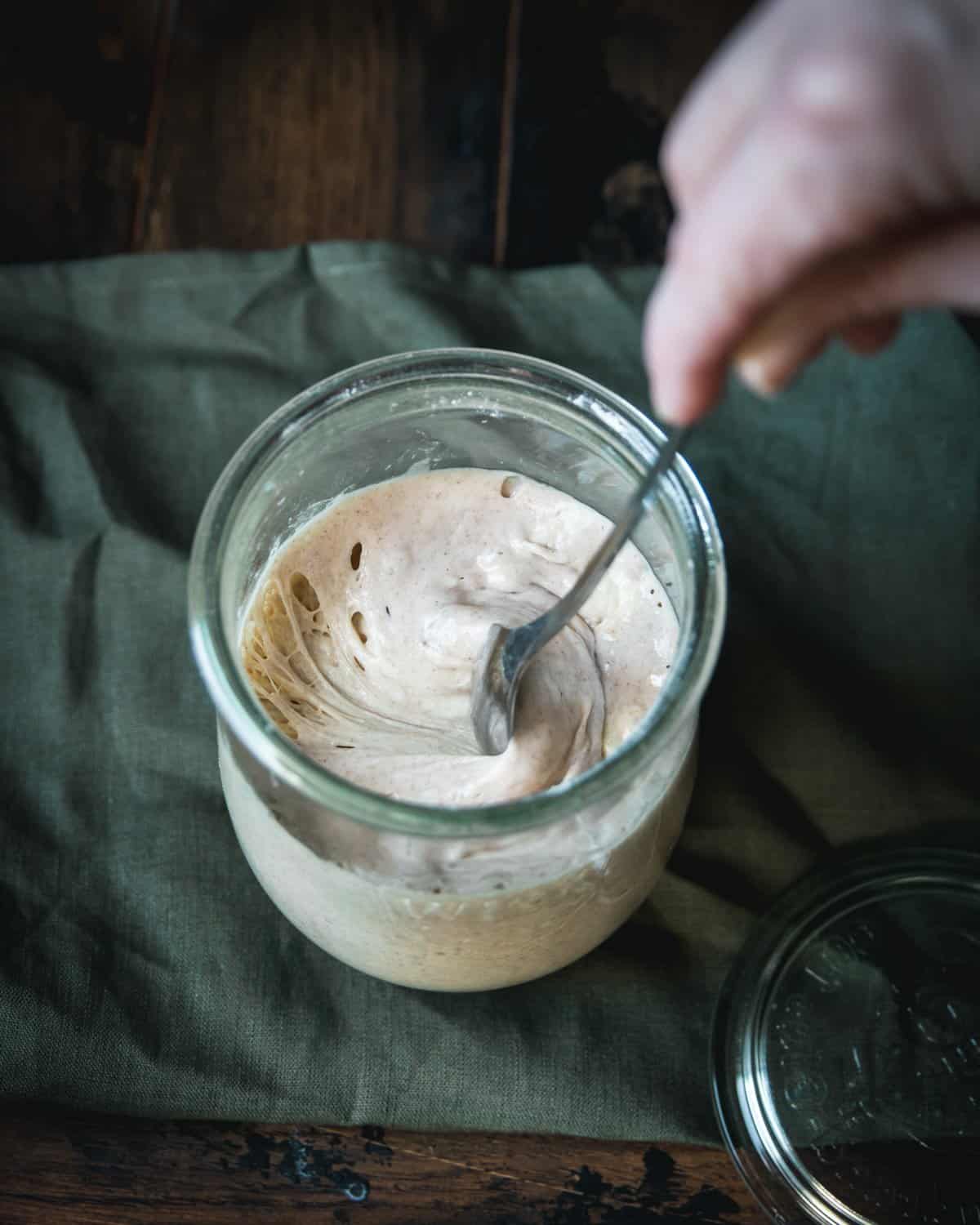 Top view of sourdough starter in a jar getting stirred sitting on a dark green natural cloth.