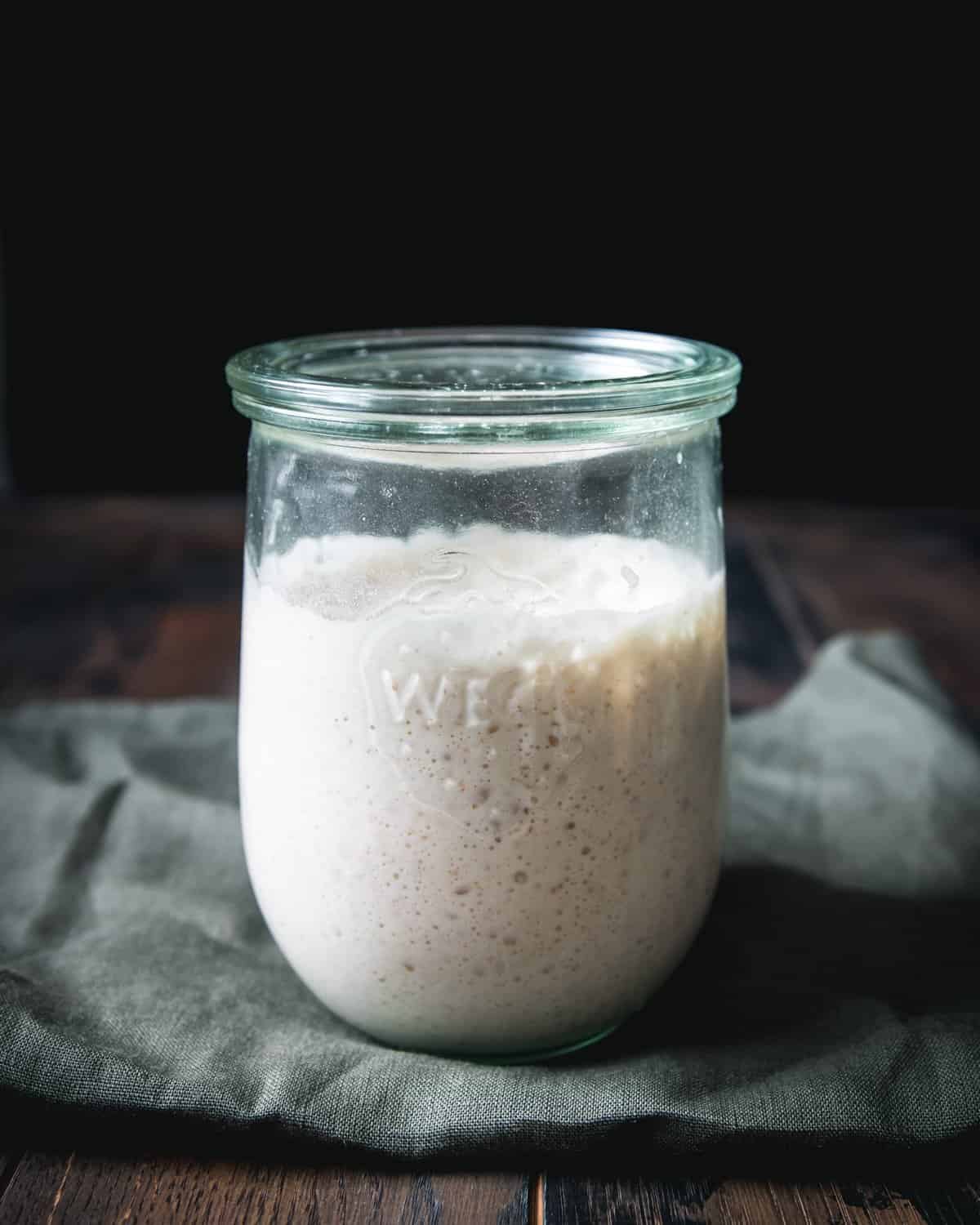 Sourdough starter in a jar with a lid on, it's bubbling away. Sitting on a dark green cloth on a dark wood background.