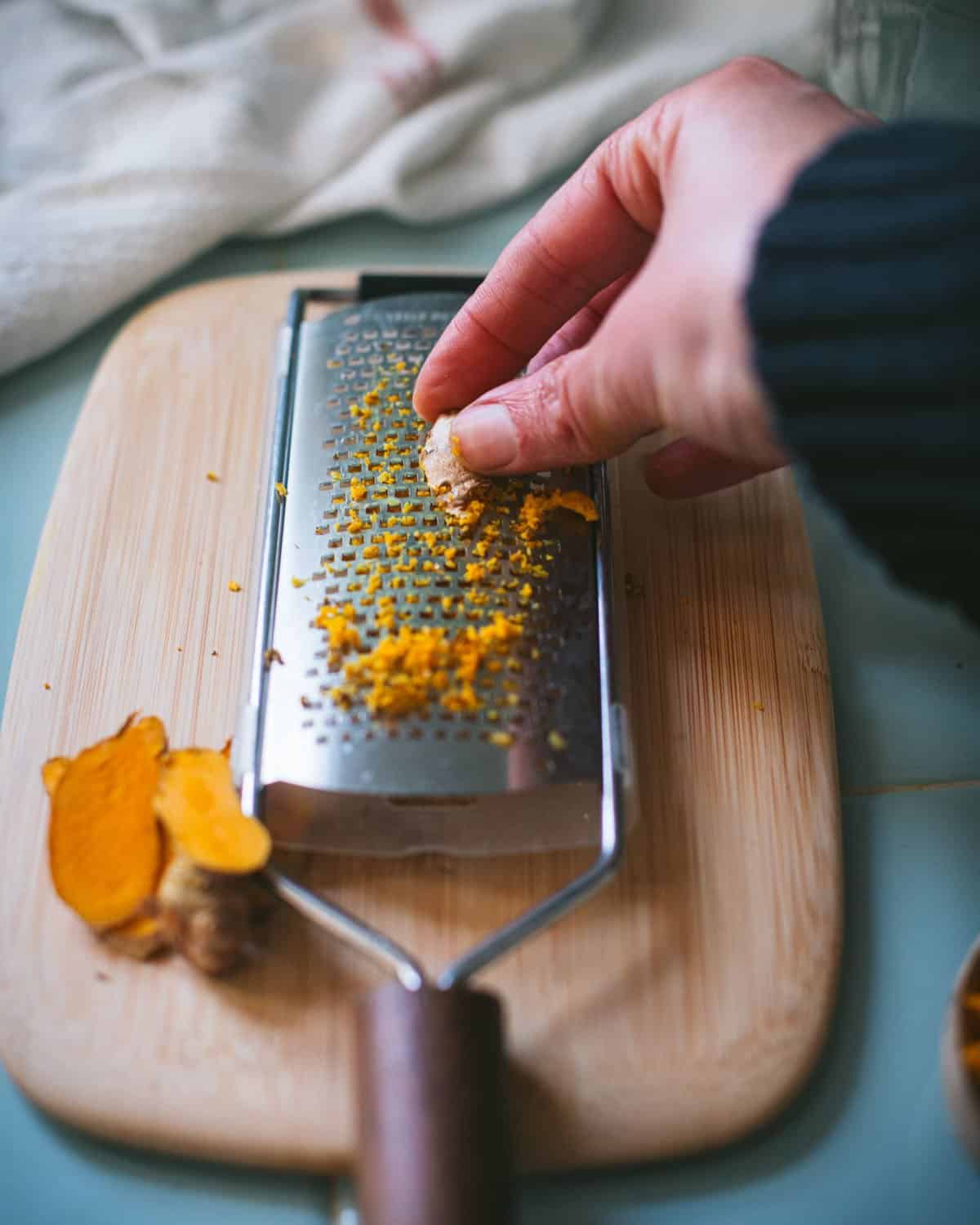 a hand grating fresh turmeric root onto a wooden cutting board