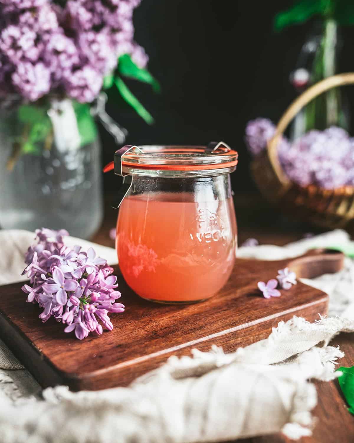 A jar of finished pink lilac jelly on a wood cutting board surrounded by fresh lilacs. 