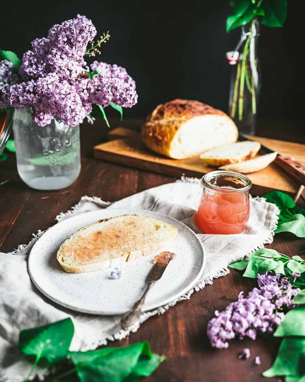 An open jar of lilac jelly sitting by a plate of toast that has lilac jelly spread on it, surrounded by fresh lilacs. 
