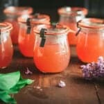 Jars of pink lilac jelly on a dark wood surface, surrounded by fresh lilacs.