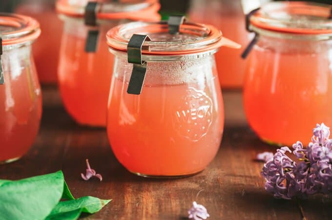 Jars of pink lilac jelly on a dark wood surface, surrounded by fresh lilacs.