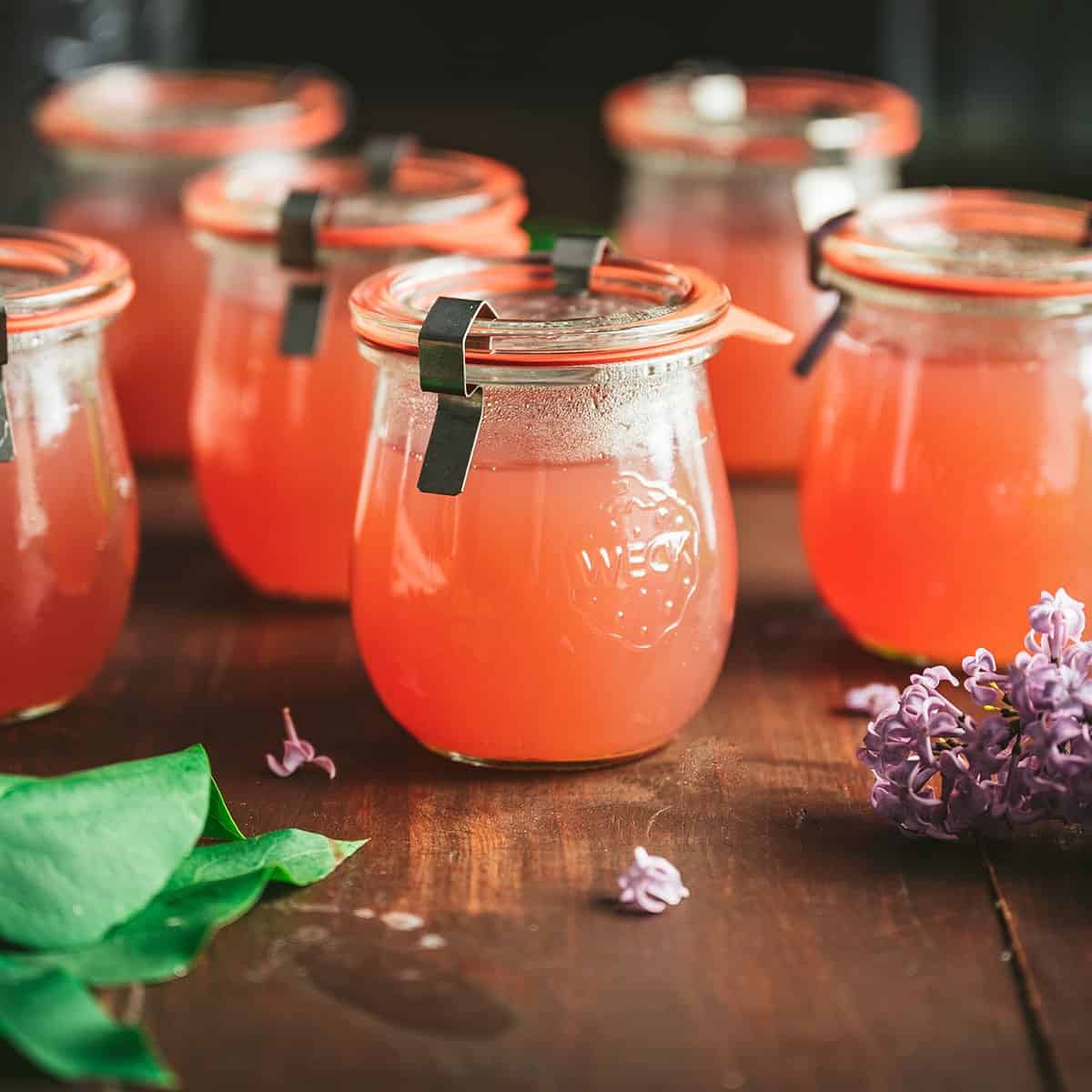 Jars of pink lilac jelly on a dark wood surface, surrounded by fresh lilacs. 